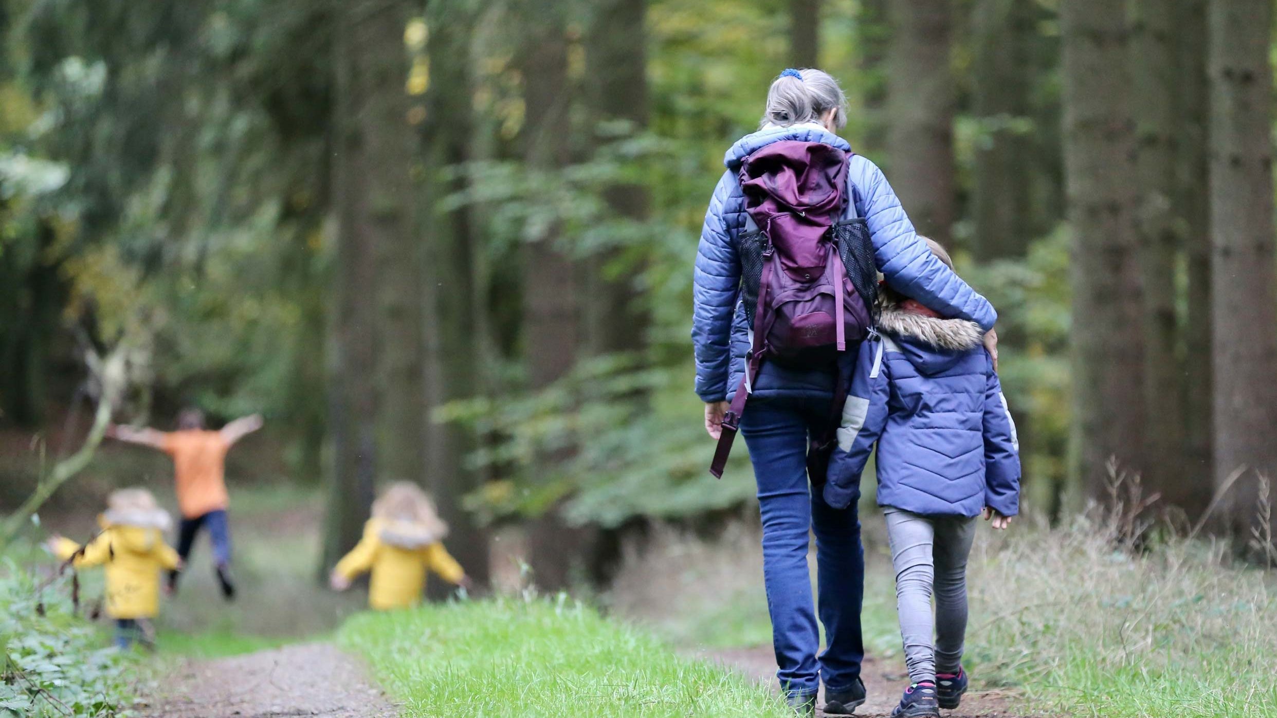 Zwei Kinder toben auf einem Waldweg, dahinter geht eine Mutter mit ihrem Arm um ein weiteres Kind.