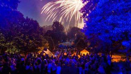 Gäste des Sommernachtsballs stehen im Freien und bewundern gemeinsam das farbenfrohe Feuerwerk am Himmel.