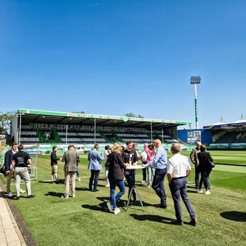 Personen an Stehtischen am Spielfeldrand, im Hintergrund die Nordtribüne des Sportparks