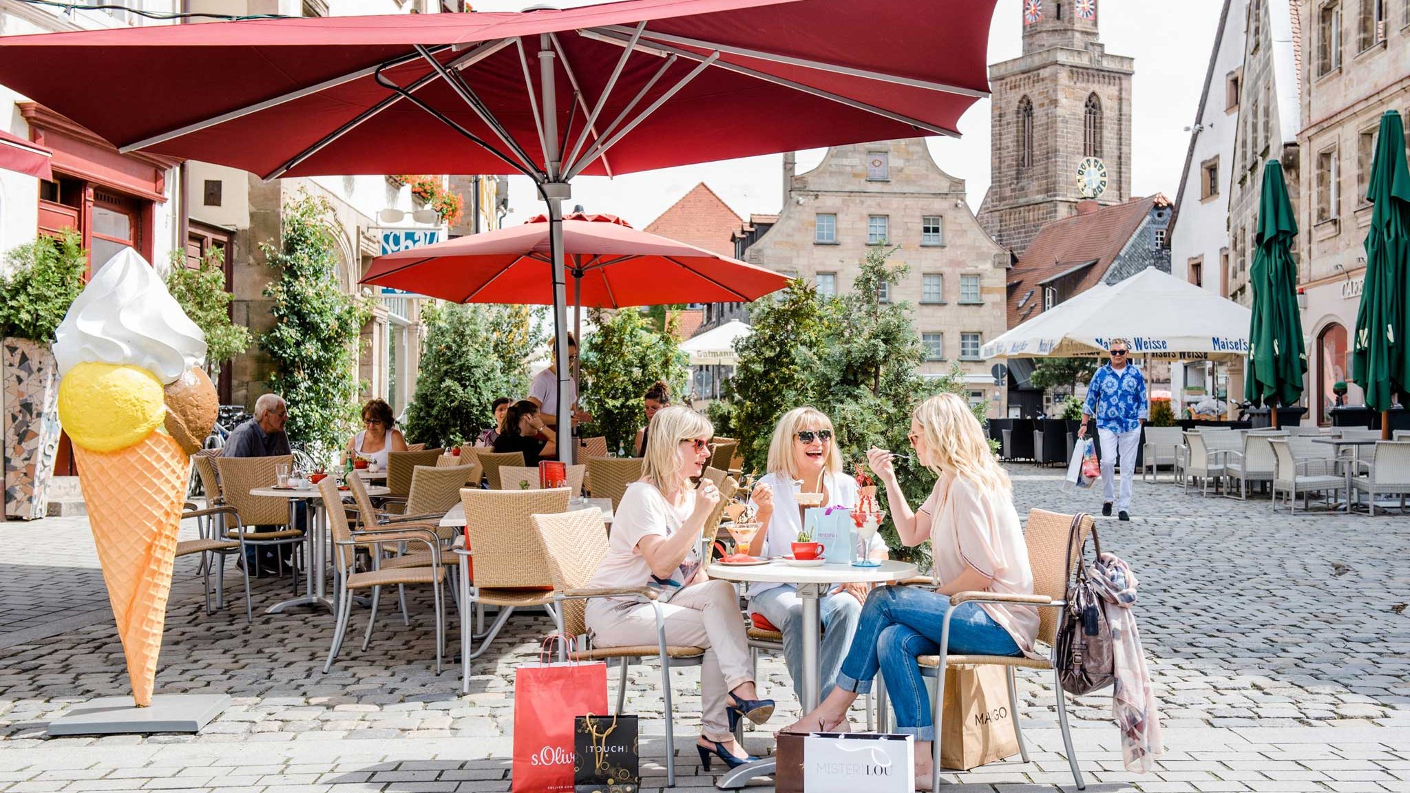 Drei Frauen genießen Eis unter einem roten Sonnenschirm am Grünen Markt mit historischen Häusern im Hintergrund