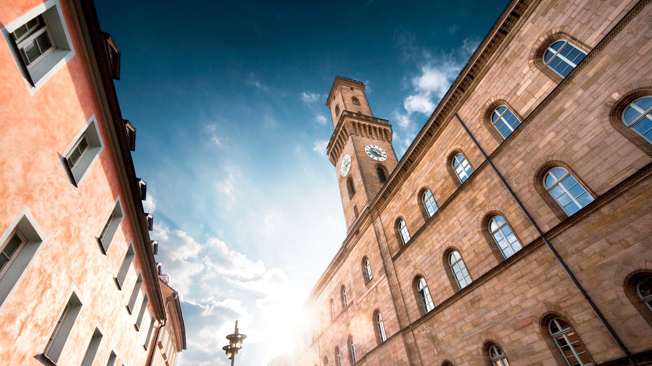 Von unten fotografierter Rathausturm mit Sonne dahinter, blauem Himmel und leichten Wolken.