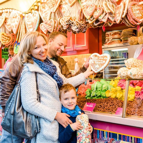 Familie steht an einem Süßwarenstand der Kirchweih mit gebrannten Nüssen, Popcorn und Lebkuchenherzen.
