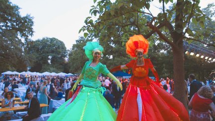 Zwei Stelzenfiguren in grünen und roten Kleidern treten vor tanzenden Sommerball-Gästen auf.