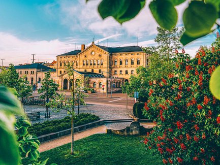 Sandsteinfassade des Fürther Hauptbahnhofs im Sonnenlicht, umrahmt von Blumen im Vordergrund