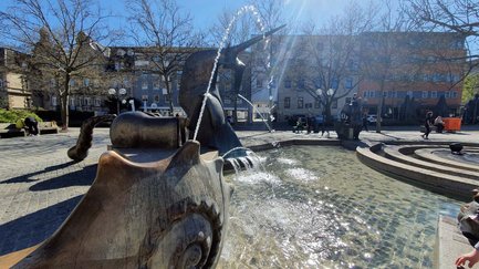 Wasserspiele einer modernen Bronzeskulptur mit Fabelwesen auf einem sonnigen, gepflasterten Platz.