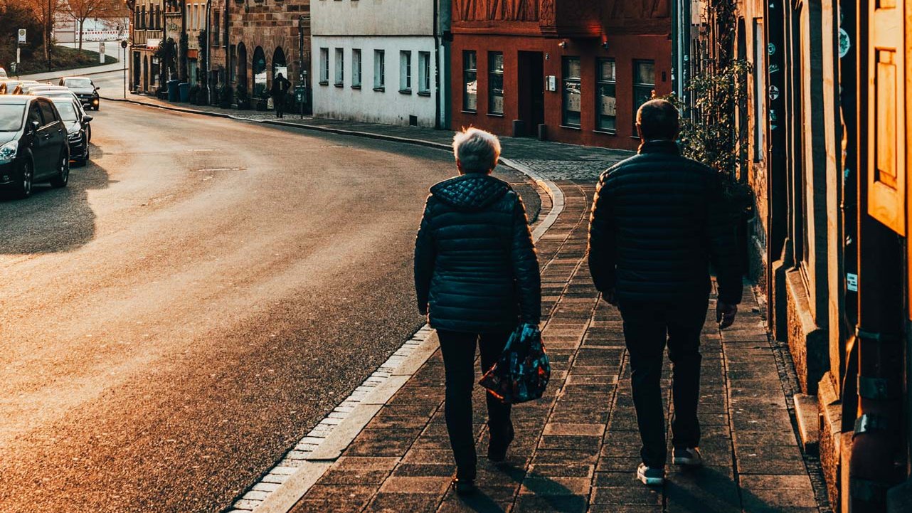 Zwei Personen laufen bei goldenem Sonnenlicht an einem roten Fachwerkhaus und Sandsteinfassaden vorbei.