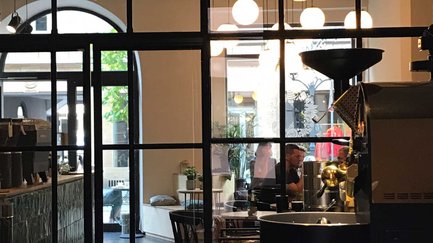 Blick durch eine Sprossenwand aus Glas auf eine Kaffeeröstmaschine und den hellen Cafébereich.