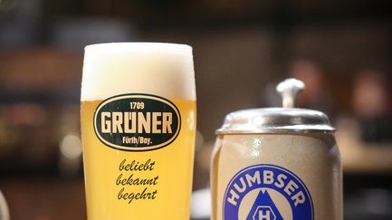 Ein gefülltes Grüner Bierglas mit Schaumkrone steht neben einem klassischen Humbser Steinkrug.