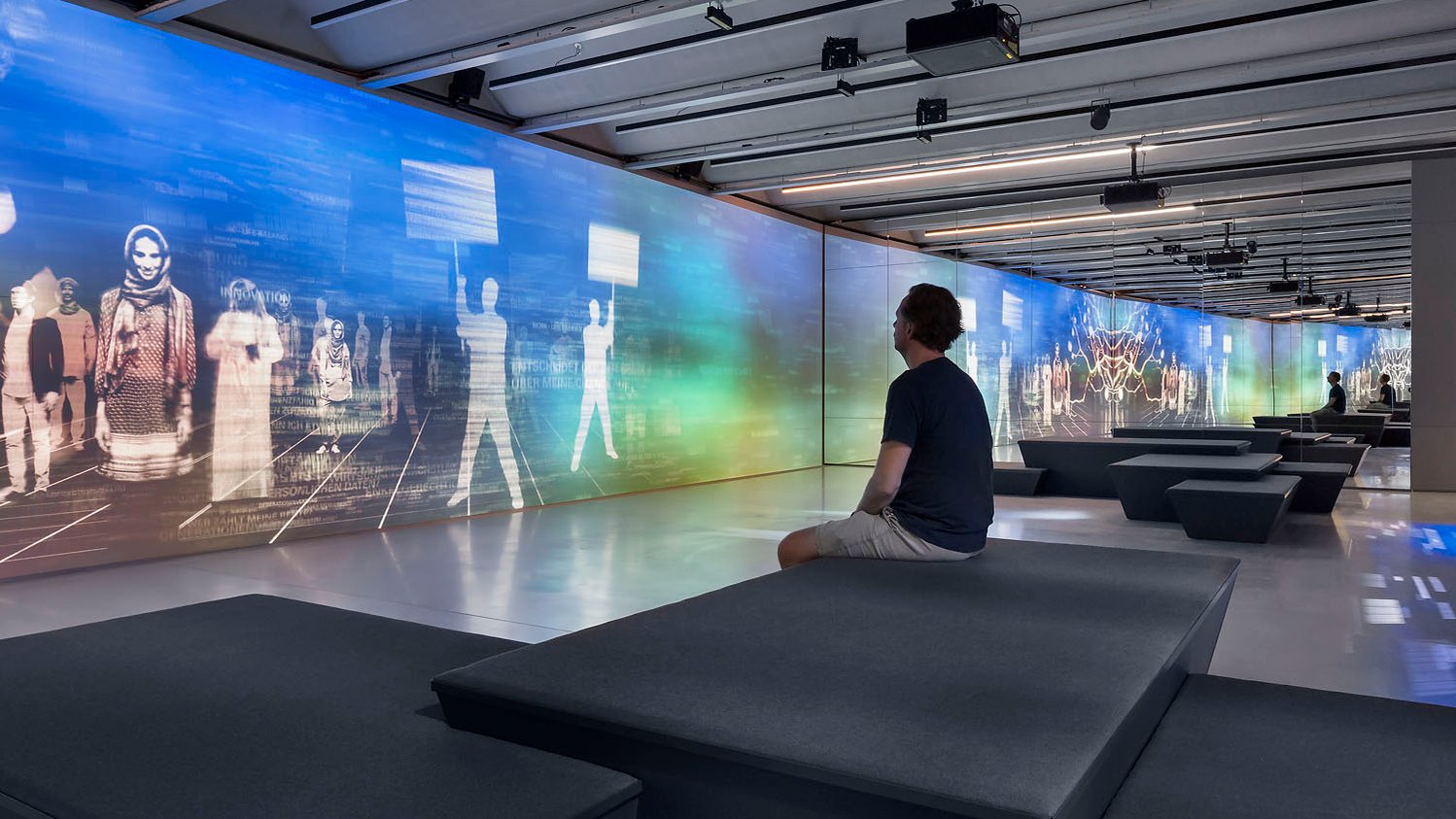 Mann sitzt im Zukunftsraum des Ludwig Erhard Zentrum auf einer Bank und betrachtet aufmerksam die Medieninstallation