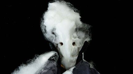 Person mit kunstvoller, watteähnlicher Maske vor dunklem Hintergrund in einer mystischen Szene.