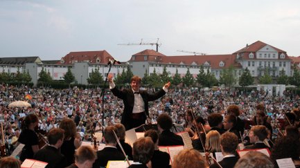 Auf der Bühne spielt ein Orchester mit Dirigent, während im Hintergrund die Besucher des Südstadt Klassik Open Air sitzen.