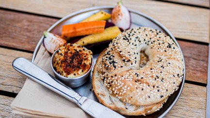 Ein Sesam-Bagel auf einem Metallteller mit Hummus, Gemüsesticks und Feigen auf einem Holztisch.