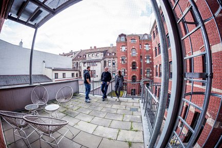 Drei Personen stehen auf einer Terrasse, im Hintergrund Backsteinhäuser