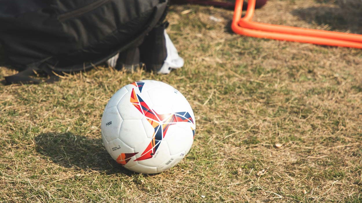 Fußball liegt auf Wiese, im Hintergrund Trainingsmaterial und Sporttaschen im Freien.