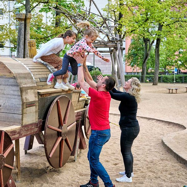 Eine Familie spielt an einer großen Holz-Eisenbahn auf einem Sandspielplatz in einer Parkanlage.