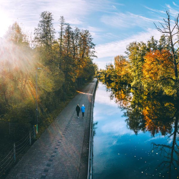 Zwei Personen spazieren an einer Uferpromenade mit bunten Bäumen, die sich im Wasser spiegeln