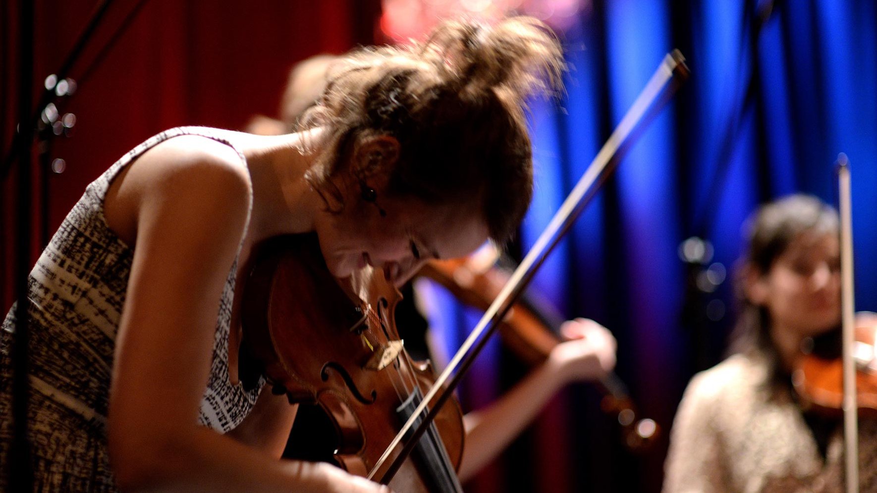Frau spielt Geige lächelnd in kleinem Ensemble, blau-rote Beleuchtung, weitere Musiker unscharf