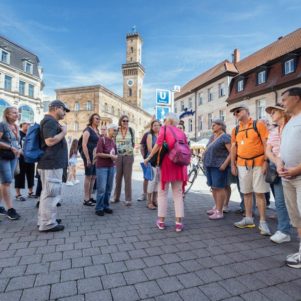 Eine Gruppe von Menschen hört bei Sonnenschein vor dem Fürther Rathausturm einer Stadtführerin zu.