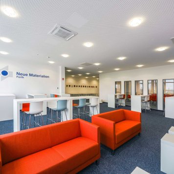 Großer Raum bei Neue Materialien Fürth mit Stehtischen, hohen Stühlen, Tageslicht und Logo an der Wand