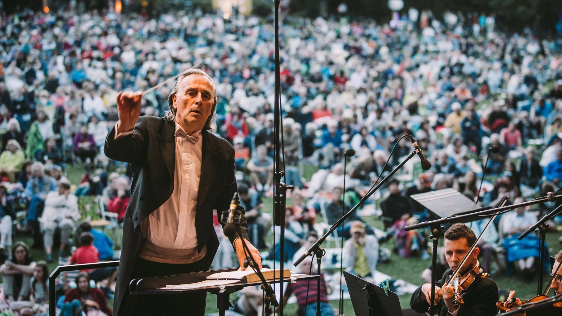 Dirigent am Pult dirigiert Orchester, Publikum sitzt auf Picknickdecken und Campingstühlen im Freien