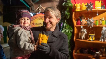 Ein Vater hält sein lachendes Kind und reicht ihm eine goldene Filzkrone an einem Stand der Altstadtweihnacht.