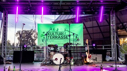 Beleuchtete Bühne mit Musikinstrumenten, Mikrofonen und grünem Kulturterrasse-Banner im Hintergrund.