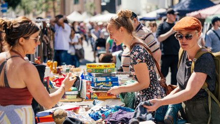 Menschen stöbern an einem bunten Flohmarktstand mit Kleidung, Spielen und Trödel im Sonnenschein.