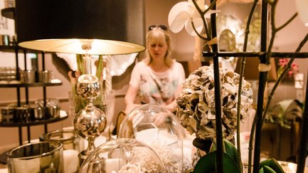 Frau schaut Wohnaccessoires und Kleinmöbel in einem Geschäft in Fürth an, leuchtende Lampe und Kunstblume im Vordergrund
