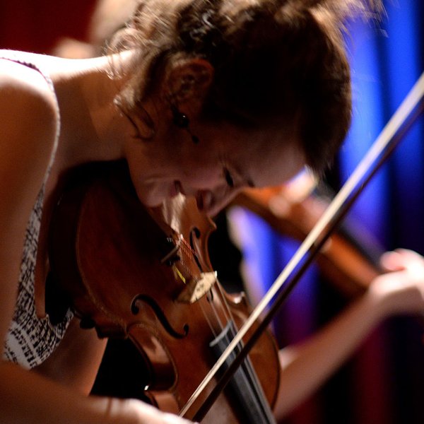 Geigerin spielt leidenschaftlich, beugt sich über ihr Instrument; im Hintergrund zwei weitere Geiger