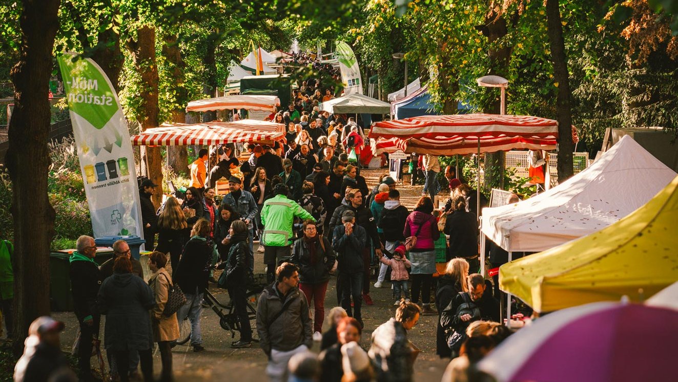 Apfelmarkt mit Marktständen entlang der Stadtpark-Allee, Besucher schlendern unter grünen Bäumen.