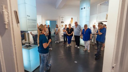 Eine Gruppe lauscht aufmerksam einer Museumsführerin vor beleuchteten Vitrinen im Museum.