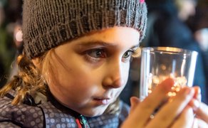 Ein kleines Mädchen hält ein Glas mit Teelicht vor ihr Gesicht und betrachtet fasziniert das warme Licht.