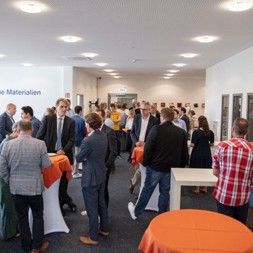 Voller Raum bei Neue Materialien Fürth mit Menschen, die an Stehtischen networken, Logo sichtbar