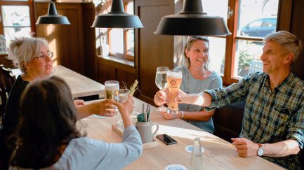 Vier Personen sitzen an einem Holztisch im Restaurant und stoßen mit Bier und Wein an.