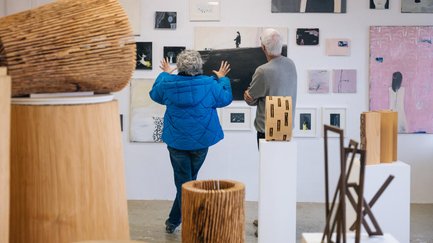 Zwei Personen betrachten schlichte Porträtkunst an weißer Wand, im Vordergrund stehen mehrere Holzskulpturen.