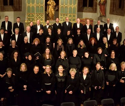 Chor Ein Kirchenchor in schwarzen Roben steht in einer Kirche, bereit für den Auftritt. Darunter sind Frauen und Männer verschiedenen Alters.