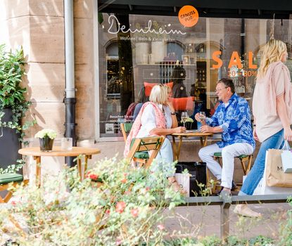 Deinheim außen Eine Frau und ein Mann sitzen vor dem Ladengeschäft Deinheim und trinken ein Getränk. Eine Frau geht an ihnen vorbei.